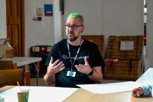 An image of a person with green hair. They are sat down next to a desk and have animated hands
