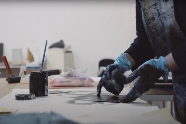 A pair of hands. They are wearing gloves and are covered in ink. Printing materials nearby.