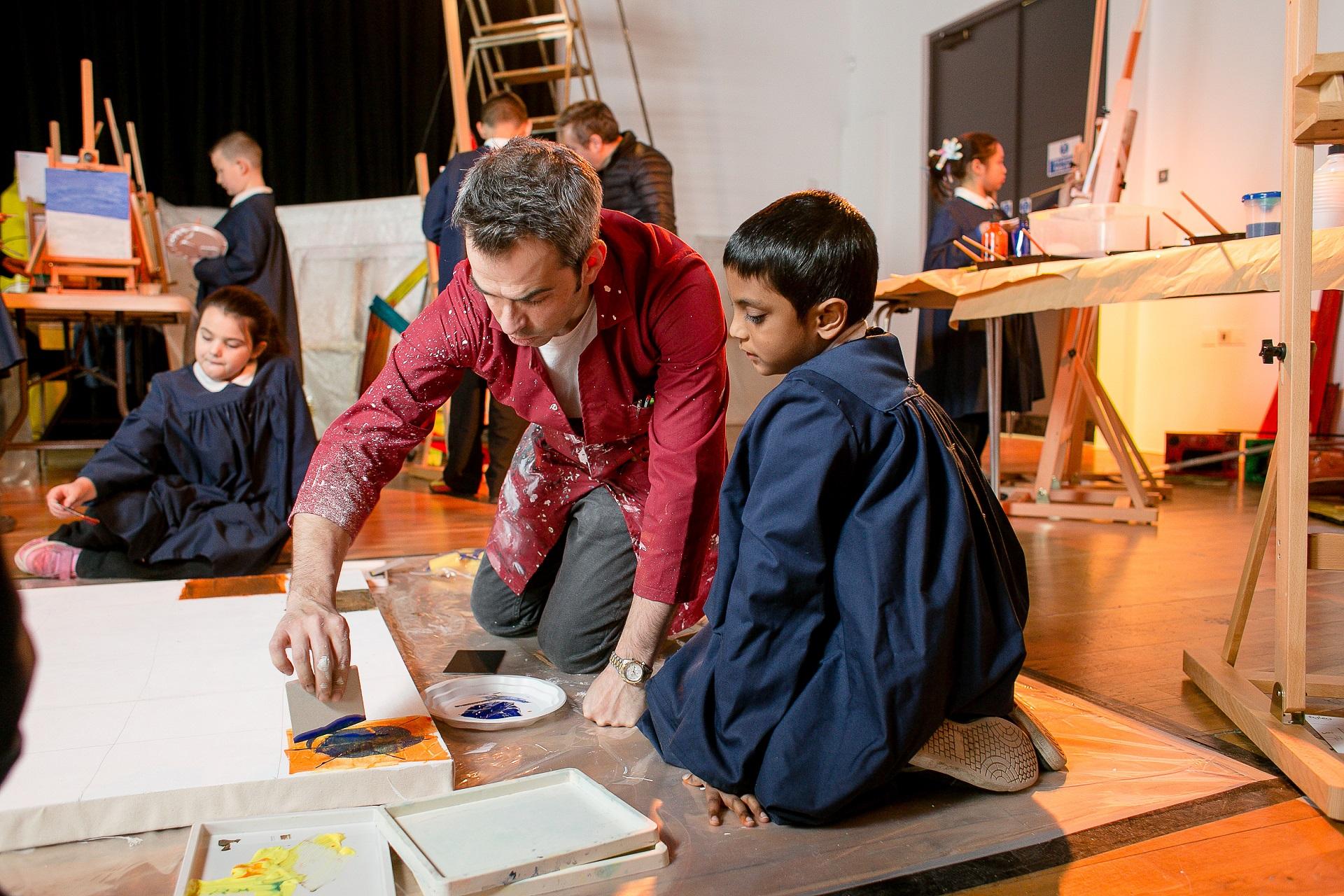 A man teaching a group of school children. They are surrounded by easels and artworks.
