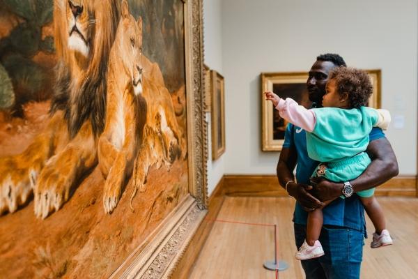 A man and young child looking at a painting of lions in a gallery