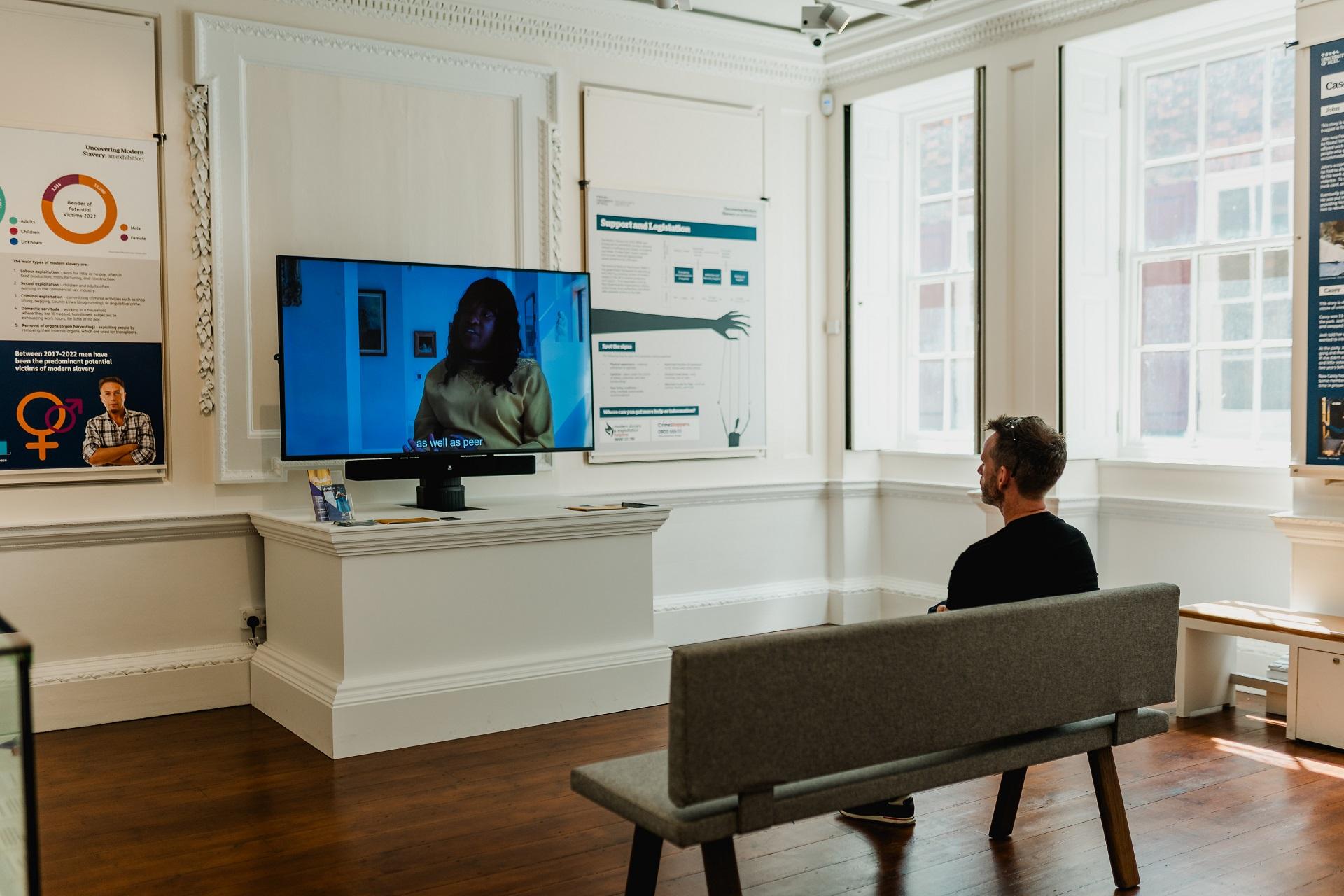 A man sat on a sofa watching a large screen tv in a museum.