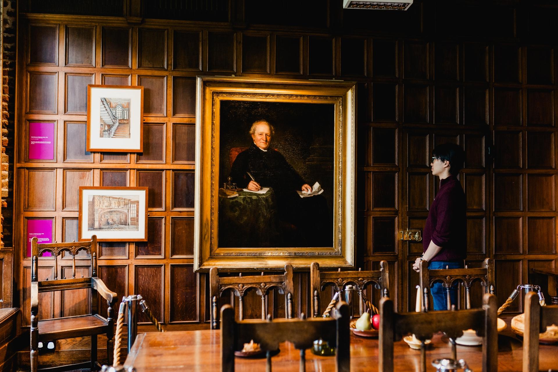 A man looking at a painting in a wooden panelled room with a wooden desk and chairs.