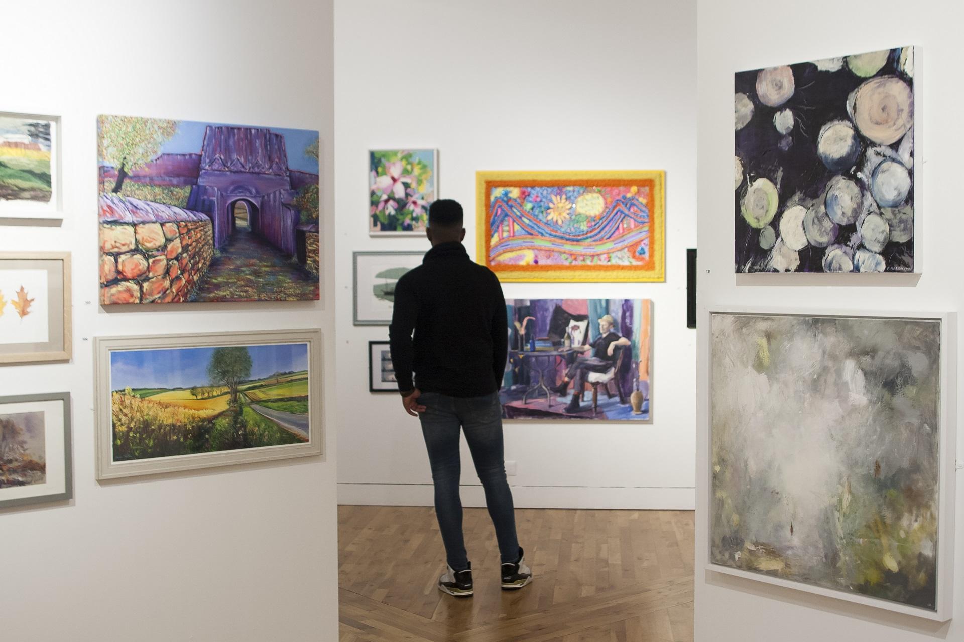 Paintings on the wall of a gallery. In the background there is a person looking at artwork.