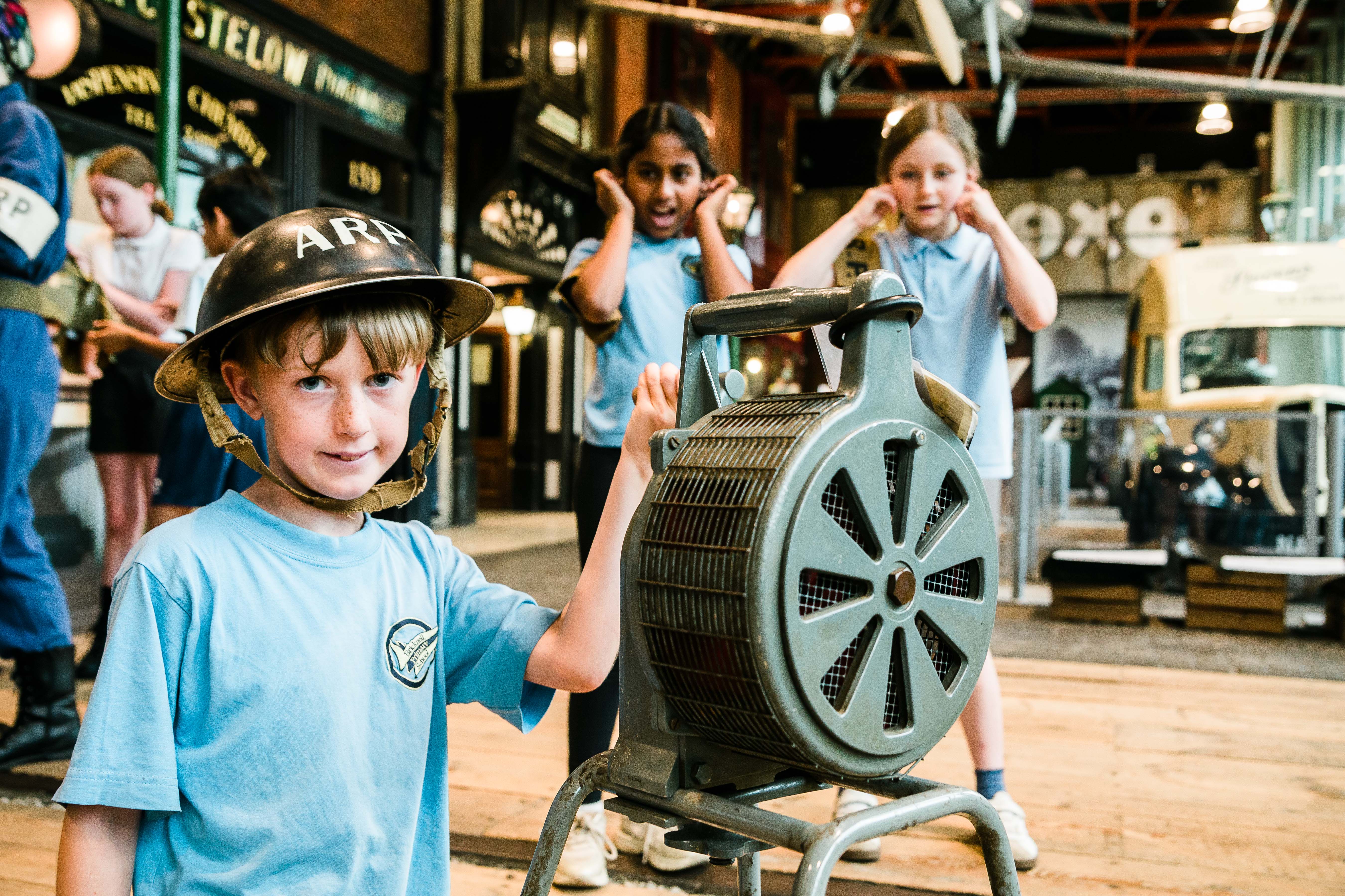 A child wearing a World War 2 ARP helmet is holding onto the handle of an air raid siren. In the background are two children with their fingers in their ears.