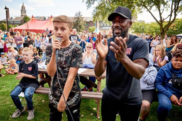A boy singing into a microphone. The person next to him is clapping.