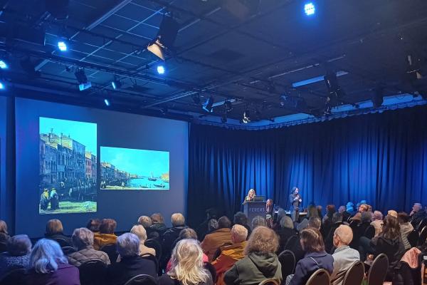 A picture of a talk at an art gallery. There are people sat in the crowd looking at a screen with paintings projected on them.