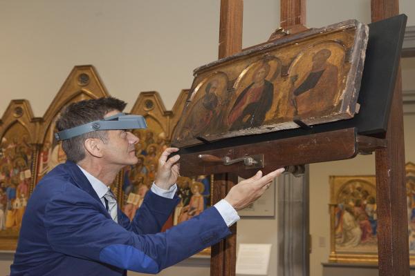A curator looking deeply at a piece of artwork. He is wearing magnified glasses and a blue lab coat