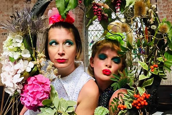 Two people in the middle of a floral wreath. They both have bright red lipstick on. One is pouting the other is looking away.