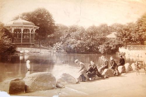 A black and white photograph of lake in a park with children sat in front of it.