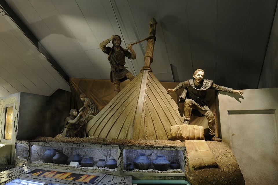 A display in a museum depicting the front of a boat with three figures stood in it.